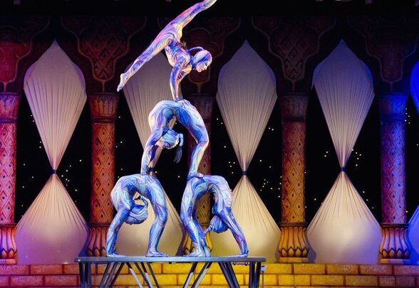 Trois acrobates en costumes irisés forment une pyramide humaine sur scène dans un décor oriental coloré - Agrandir l'image, fenêtre modale