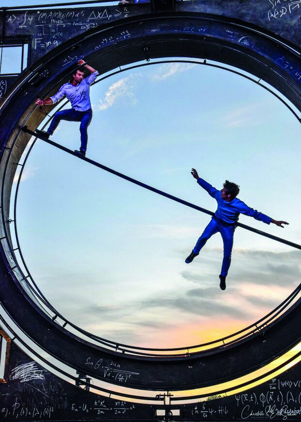 Deux interprètes dans l'Oculaire, grande roue couverte de formules mathématiques : l'un en équilibre sur une barre, l'autre suspendu dans les airs