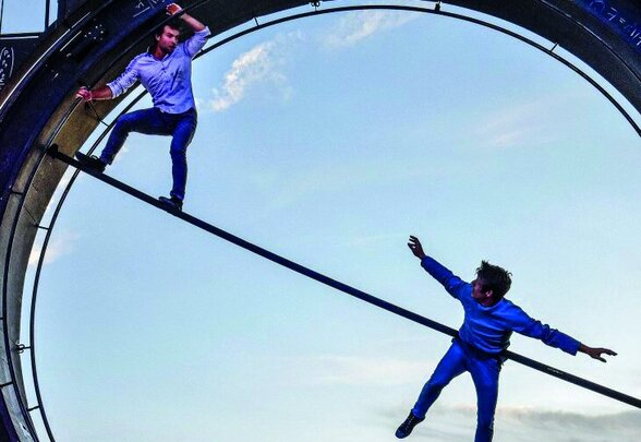Deux interprètes dans l'Oculaire, grande roue couverte de formules mathématiques : l'un en équilibre sur une barre, l'autre suspendu dans les airs - Agrandir l'image, fenêtre modale