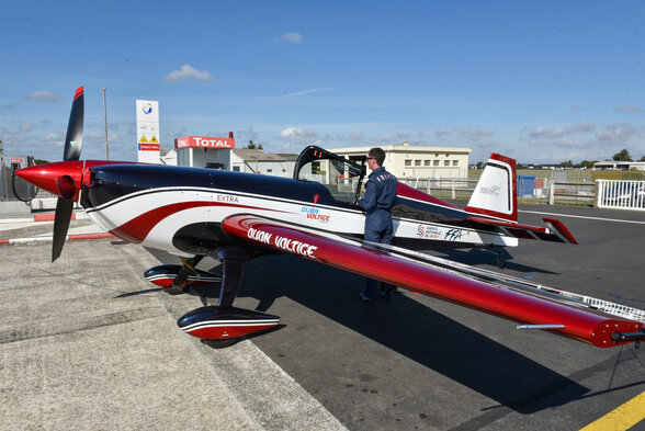 Pilote en combinaison bleue inspectant un avion de voltige aux couleurs rouges et blanches sur le tarmac. - Agrandir l'image, fenêtre modale