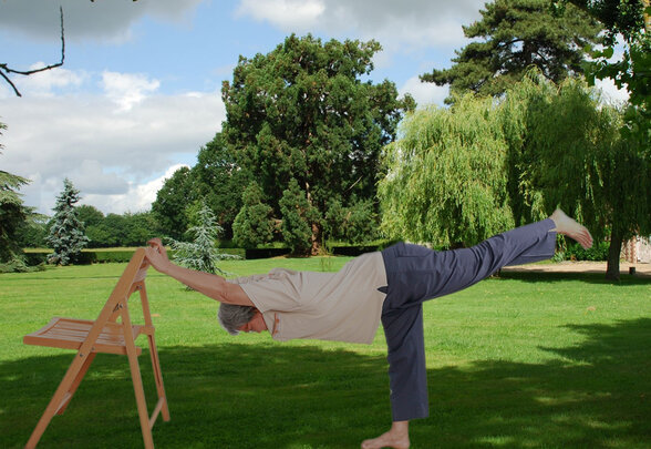 Personne réalisant une posture de yoga en équilibre sur une jambe, s'appuyant sur une chaise en bois, dans un parc verdoyant - Agrandir l'image, fenêtre modale
