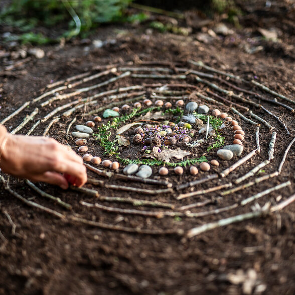 Mandala éphémère de bâtons, galets, glands et fougères en cercles concentriques sur la terre, une main le façonnant - Agrandir l'image, fenêtre modale