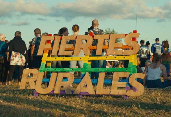 Panneau en bois « Fiertés rurales » aux couleurs arc-en-ciel, entouré de participants rassemblés dans un champ au coucher du soleil - Agrandir l'image, fenêtre modale