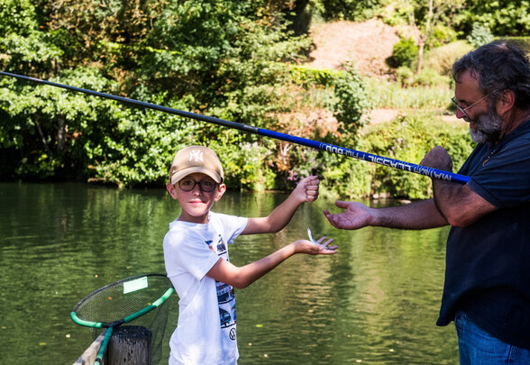 Un enfant montre un petit poisson pêché à un adulte au bord d'une rivière, canne à pêche en main - Agrandir l'image, fenêtre modale